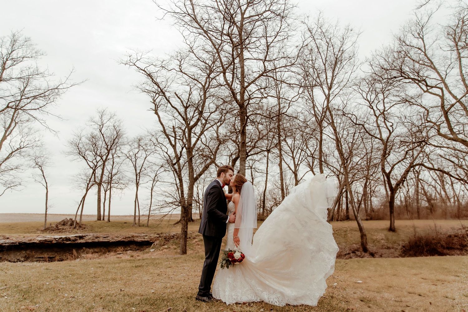 A couple embraces in a romantic moment as the bride's veil flows dramatically among bare winter trees.