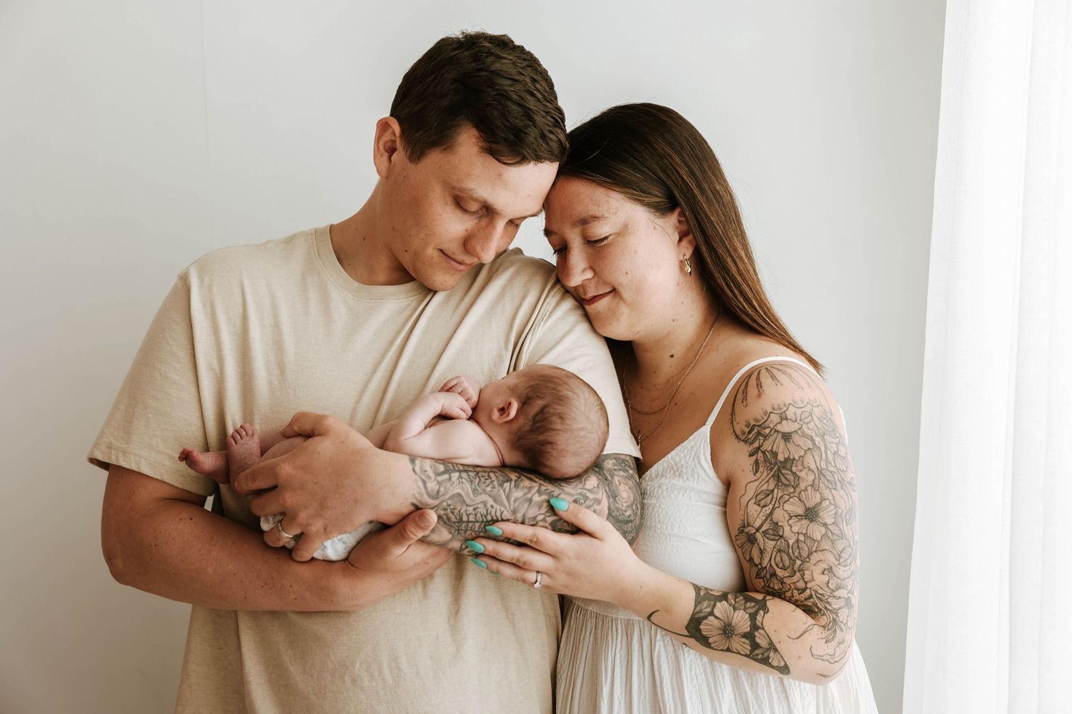 Parents in neutral tones holding and gazing lovingly at their newborn baby by a bright window.