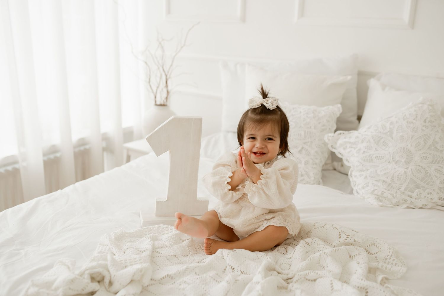 A small child in a white ruffled dress sits on a white bed with a large number one decoration for their first birthday.