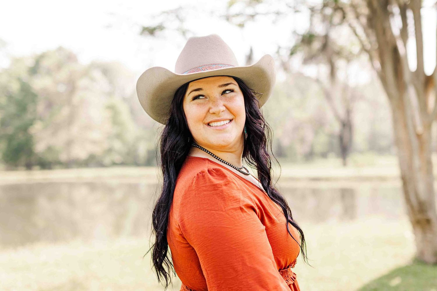 A woman in a bright orange sweater and cowboy hat smiles outdoors against a natural landscape backdrop.