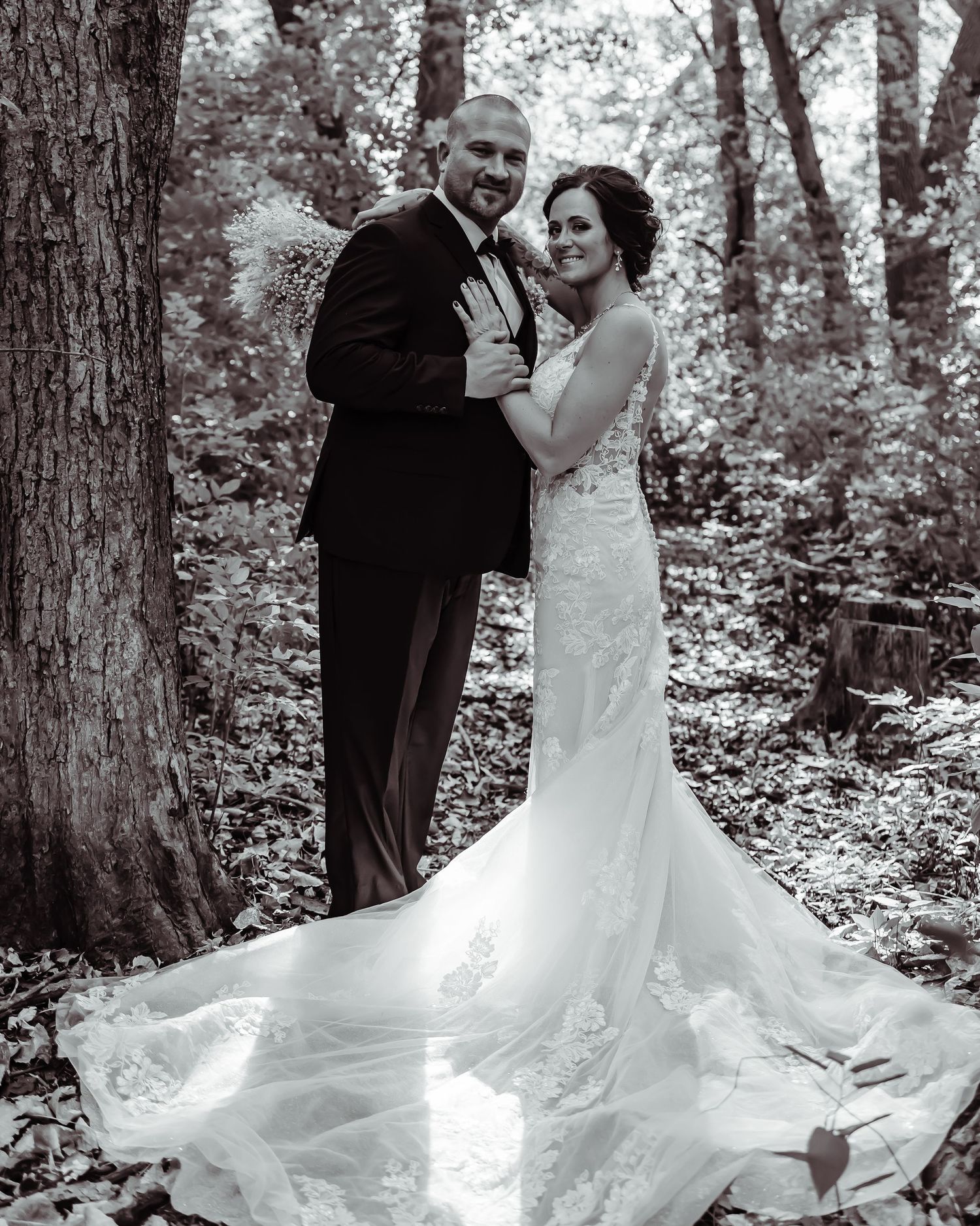 A romantic black and white wedding photo of a couple embracing in a wooded forest setting with a flowing bridal gown train.