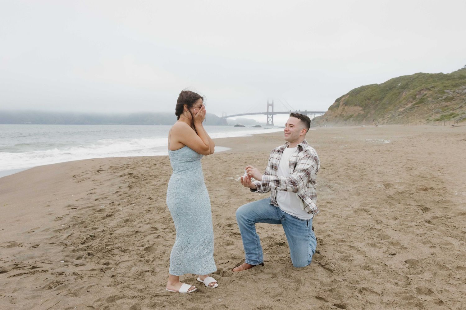 A couple stands on a foggy beach with the Golden Gate Bridge visible in the background during a special moment.