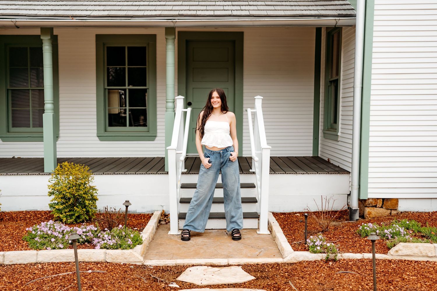 A casual figure stands on the front steps of a white farmhouse with green trim and mulched garden beds.