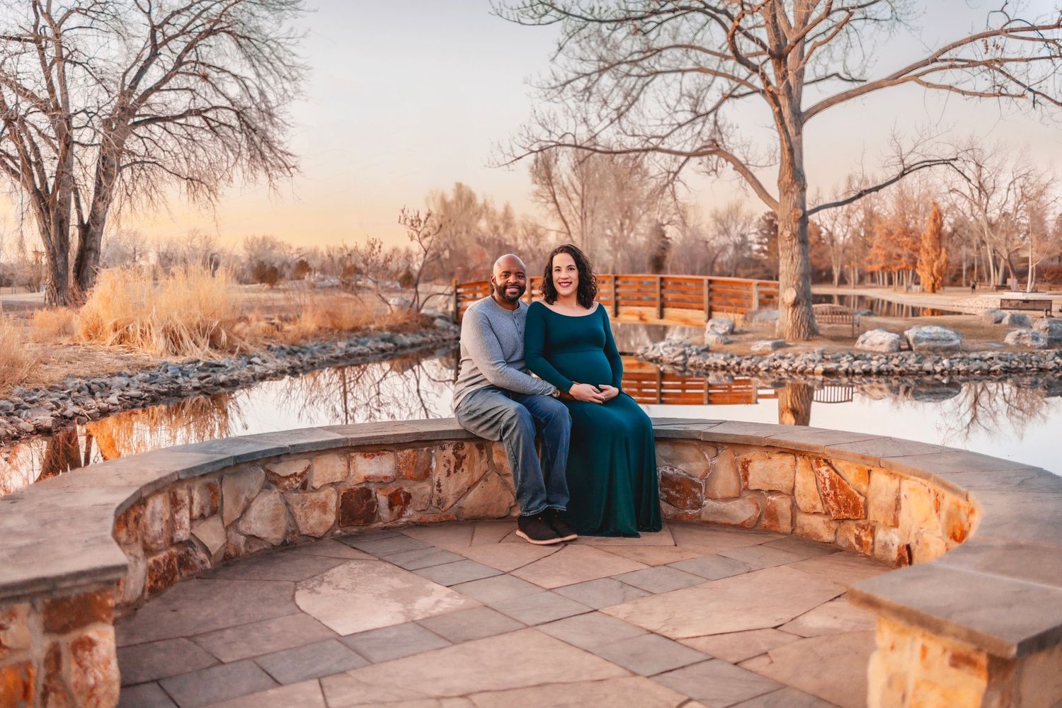 A couple sits on a curved stone bench by a frozen pond at sunset with bare winter trees in the background.  Maternity photoshoot