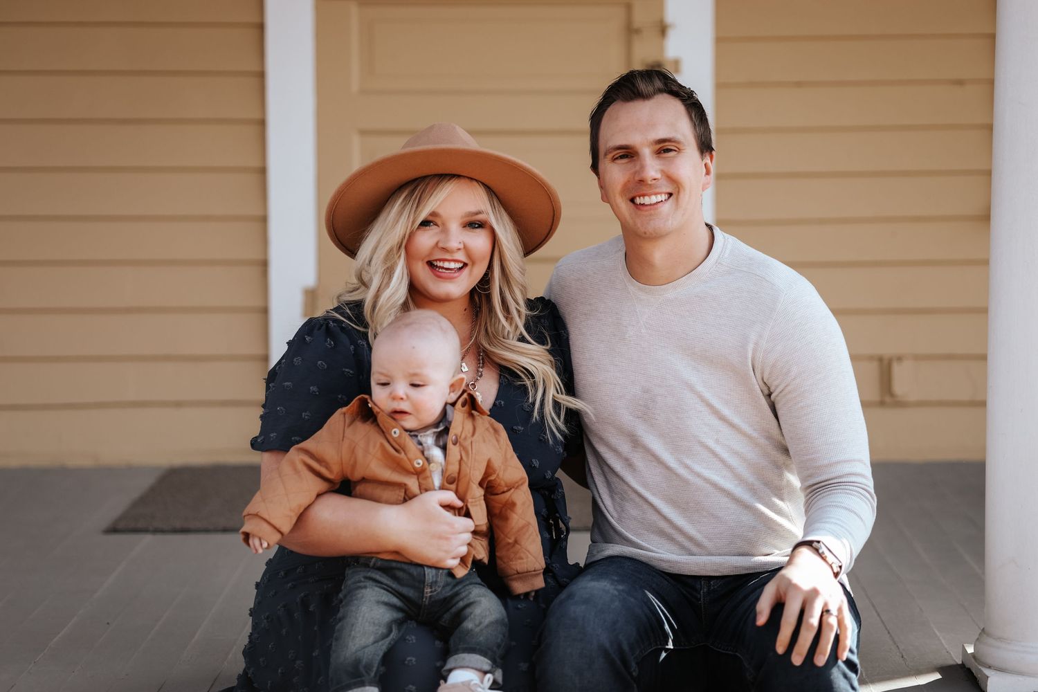 A family sits together on a porch, with the woman wearing a brown hat and the baby in casual attire.