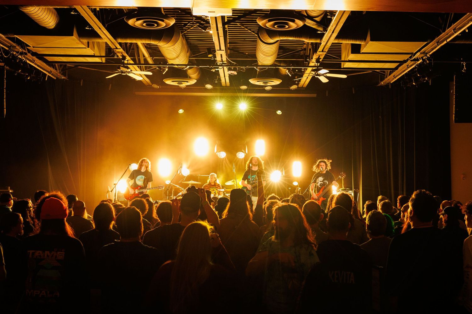 Silhouetted concert crowd watches performers on stage with dramatic orange and yellow lighting in dark venue.