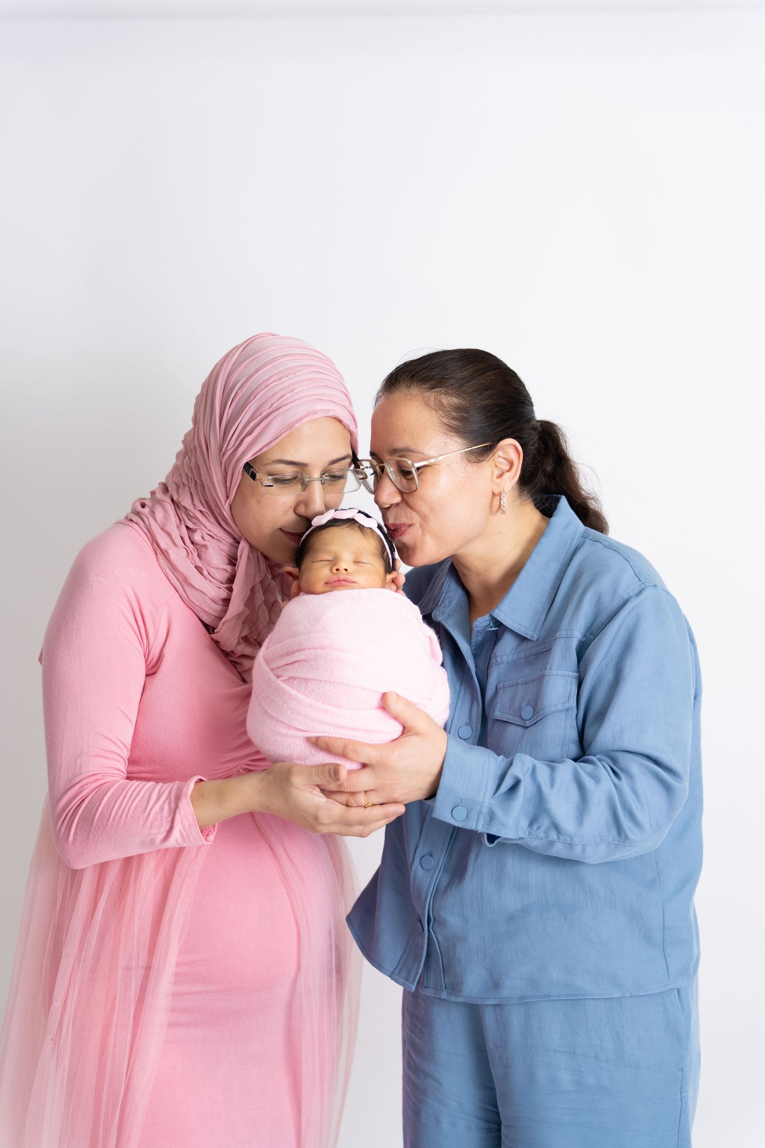 Two people in pink and blue outfits sharing a tender moment with a baby wrapped in pink blanket against white background.