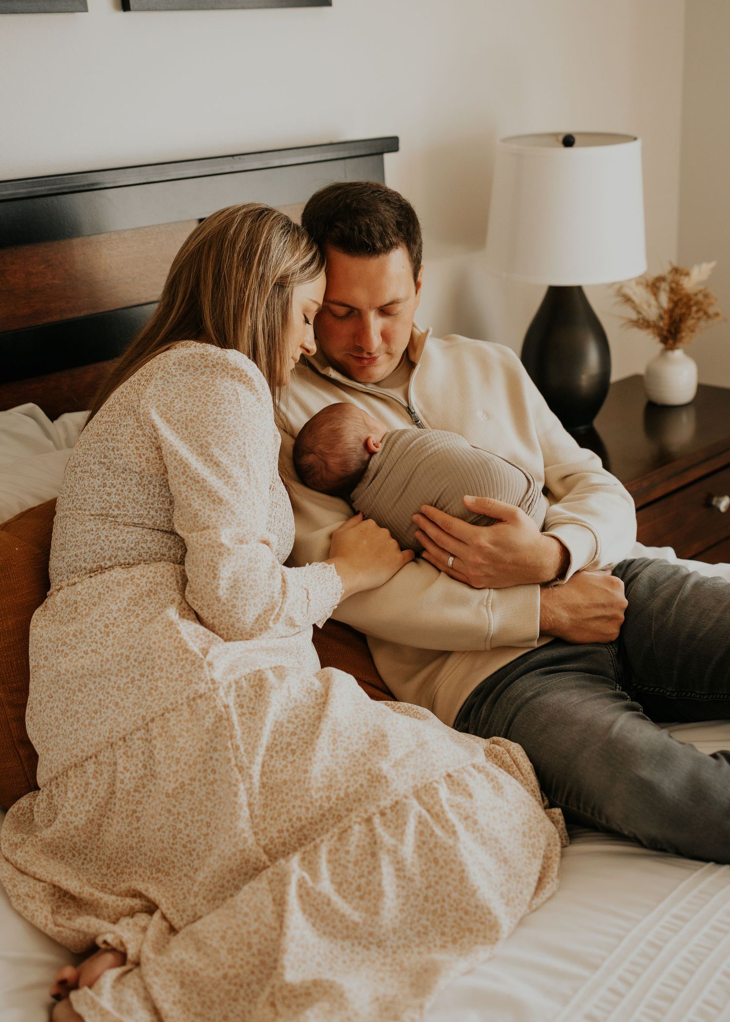 A family shares an intimate moment together on the bed with their newborn baby in soft, warm lighting.