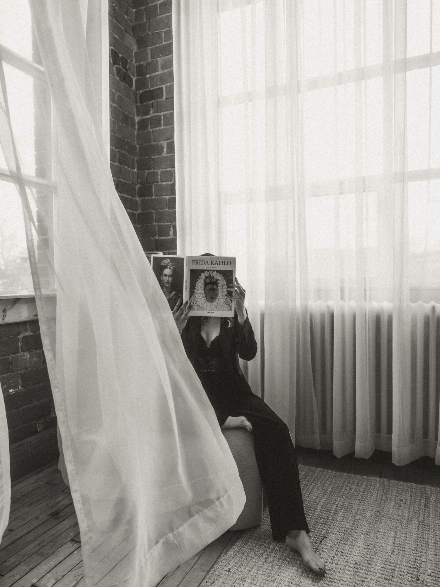 Black and white photo of someone holding a vintage camera in front of flowy white curtains by a brick wall.