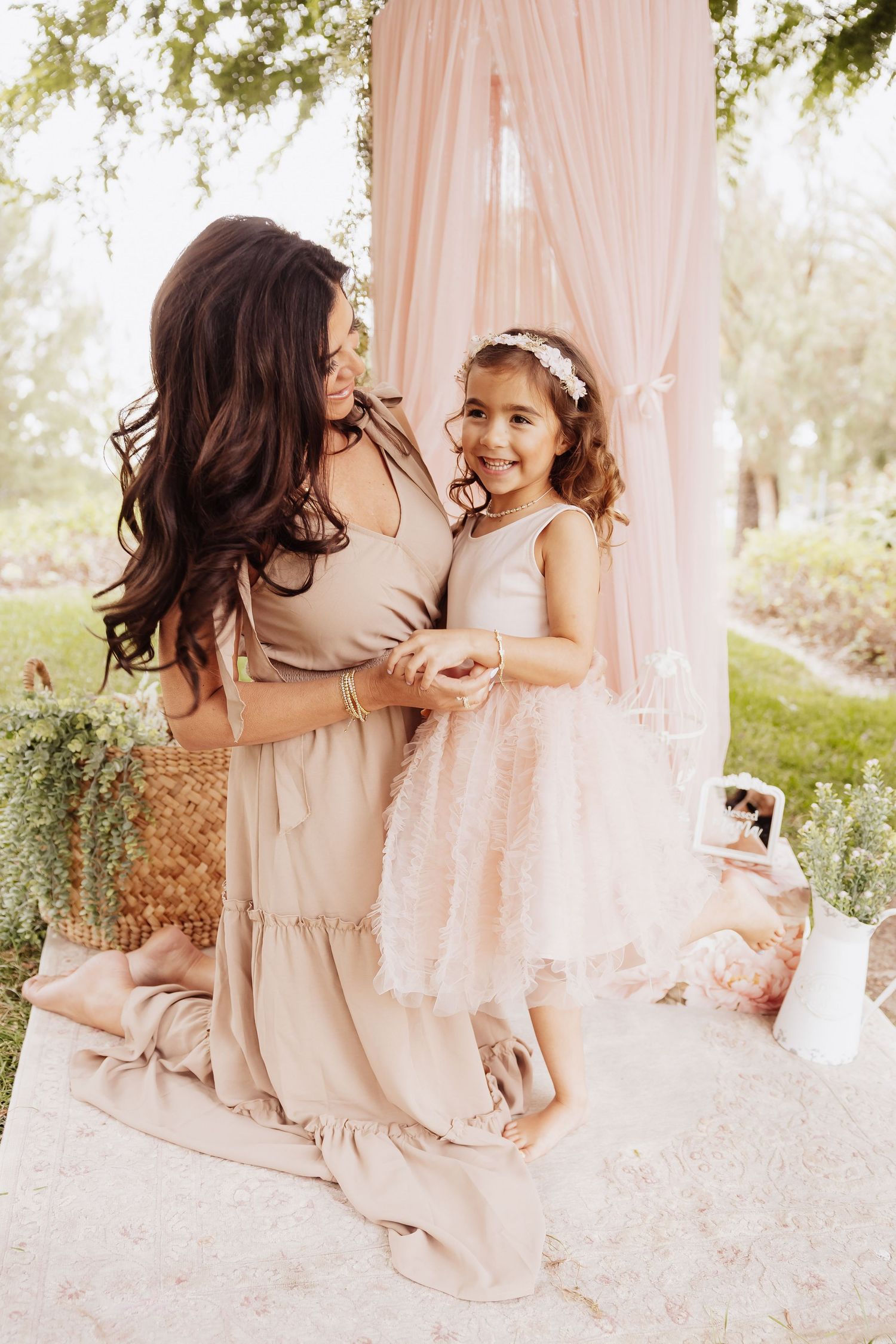 A parent and child in neutral-toned dresses share a sweet candid moment in an outdoor garden setting with flowy curtains.