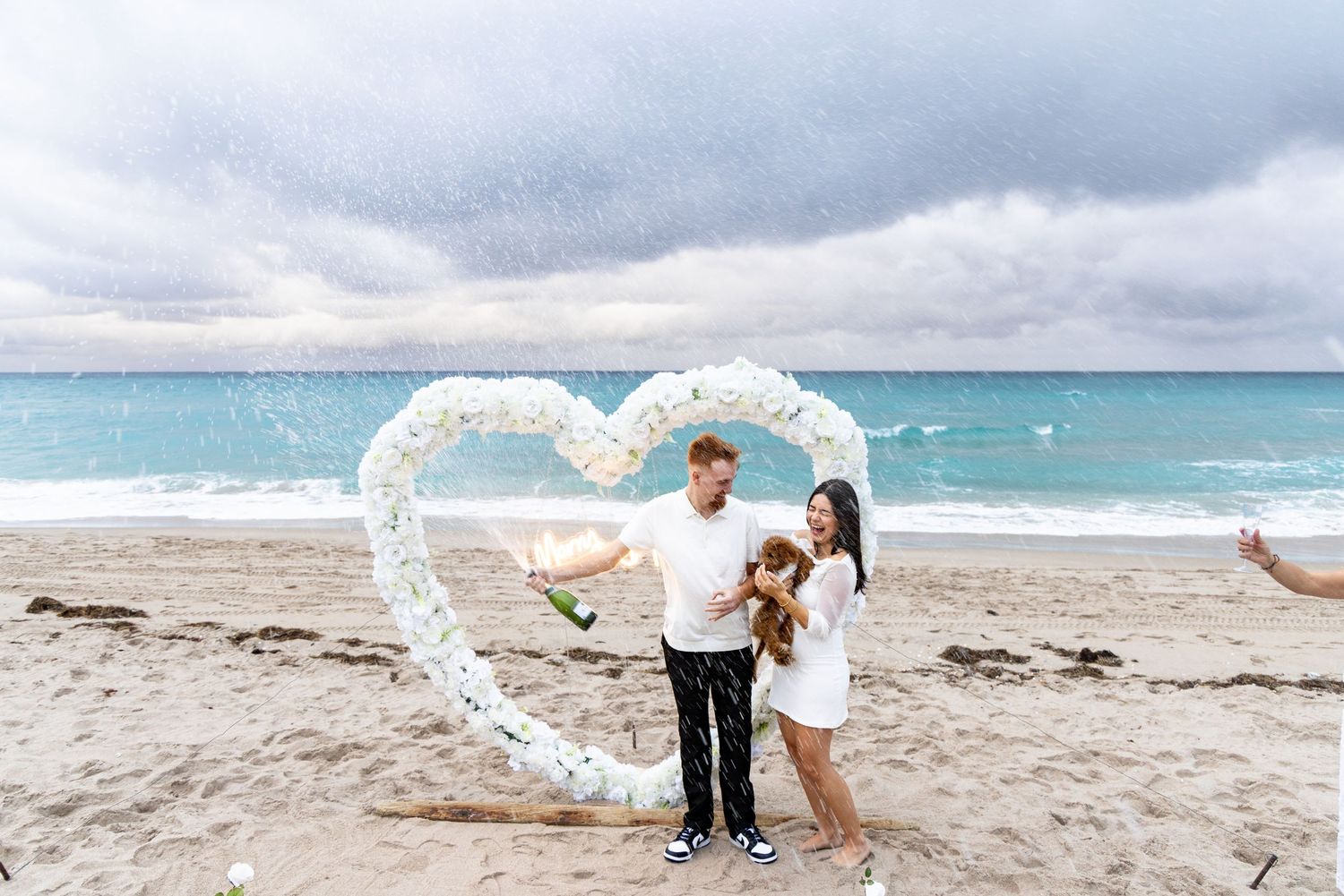 Couple posing under heart-shaped balloon arch on beach with turquoise waters.