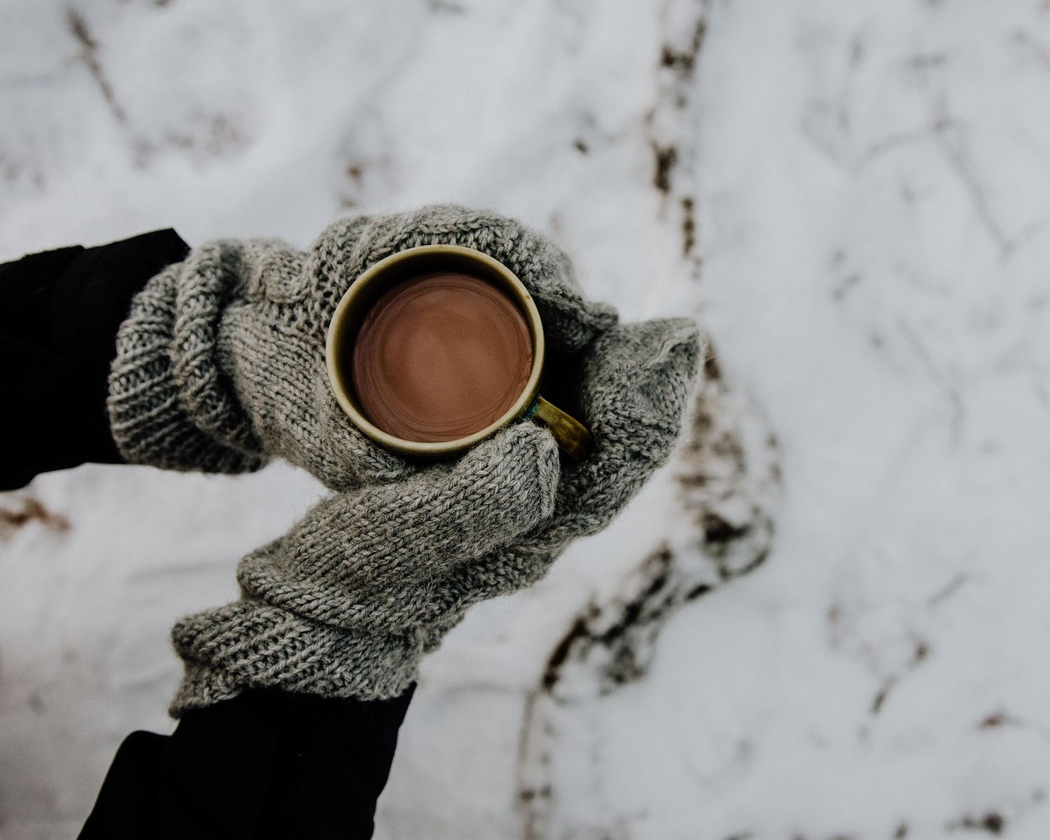 Hands wearing gray knit gloves hold a steaming cup of hot chocolate against a snowy white background.