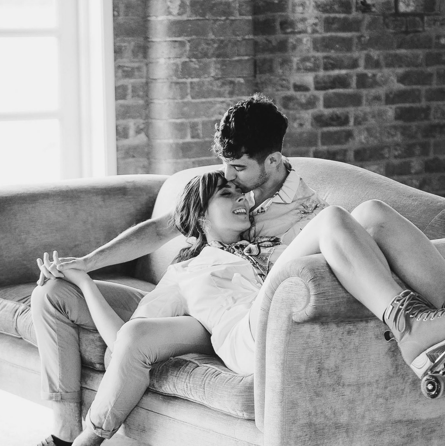 Black and white photo of a romantic couple cuddling together on a vintage leather couch against a brick wall.