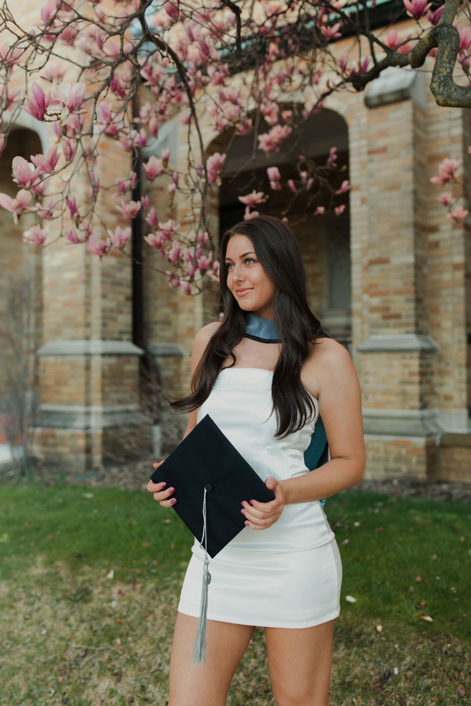 Graduate in white dress holding diploma poses under blooming pink magnolia flowers by brick building.