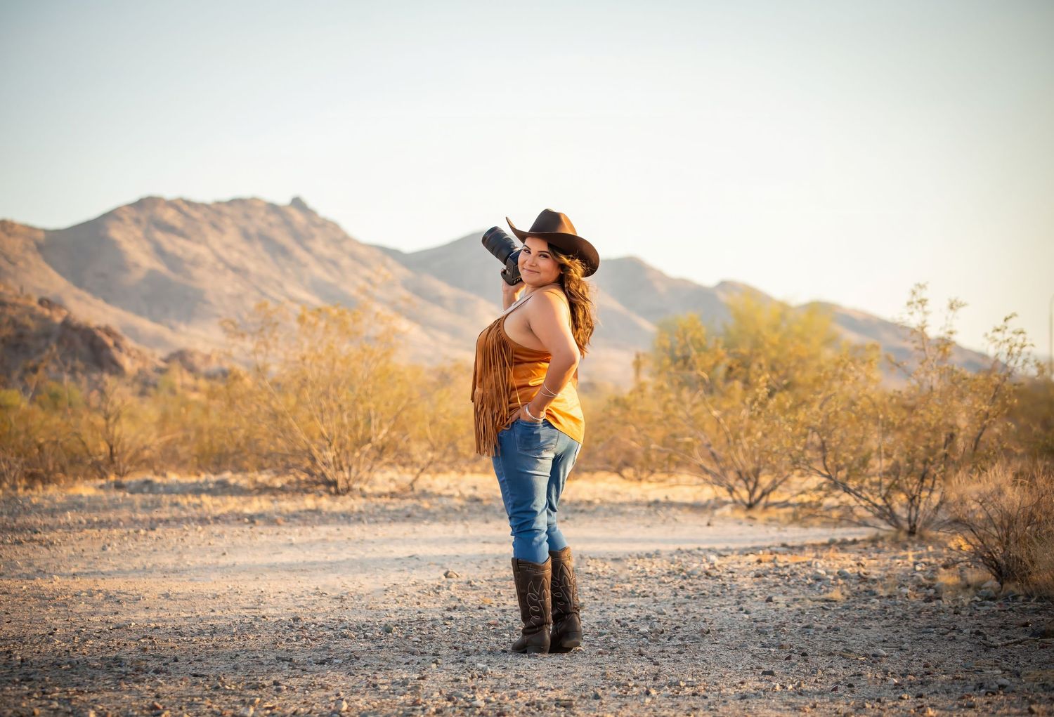 A photographer wearing jeans and boots stands in a desert landscape with mountains and golden light in the background.