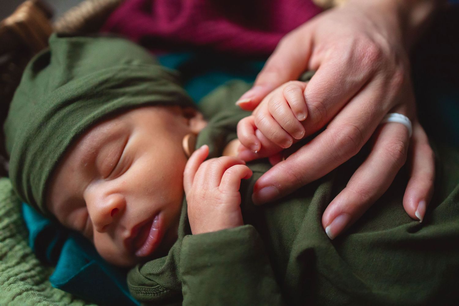 Close-up of adult hands gently holding tiny fingers of a sleeping baby wearing green clothing.