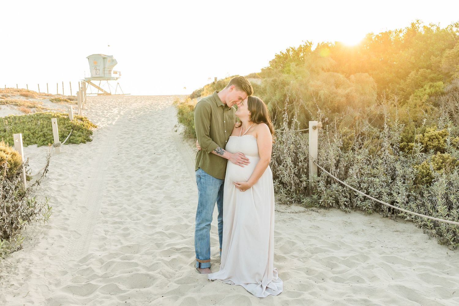 A couple shares a romantic moment on a sandy beach path during golden hour sunset, framed by coastal vegetation.