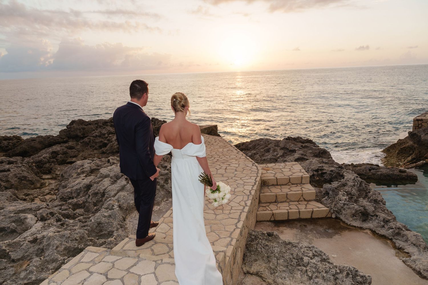 Oceanside gazebo and wooden walkway with crashing waves against rocky shoreline at sunset.
