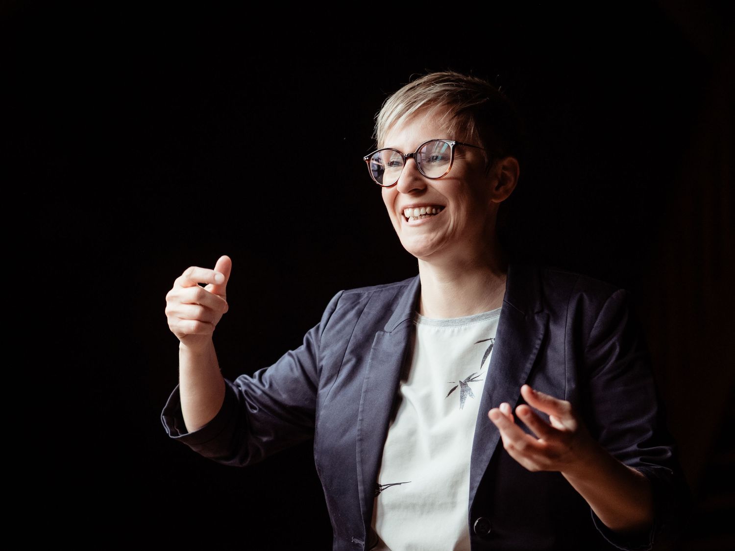 A person in a navy blazer and white shirt gestures enthusiastically while smiling and wearing glasses against a dark background.