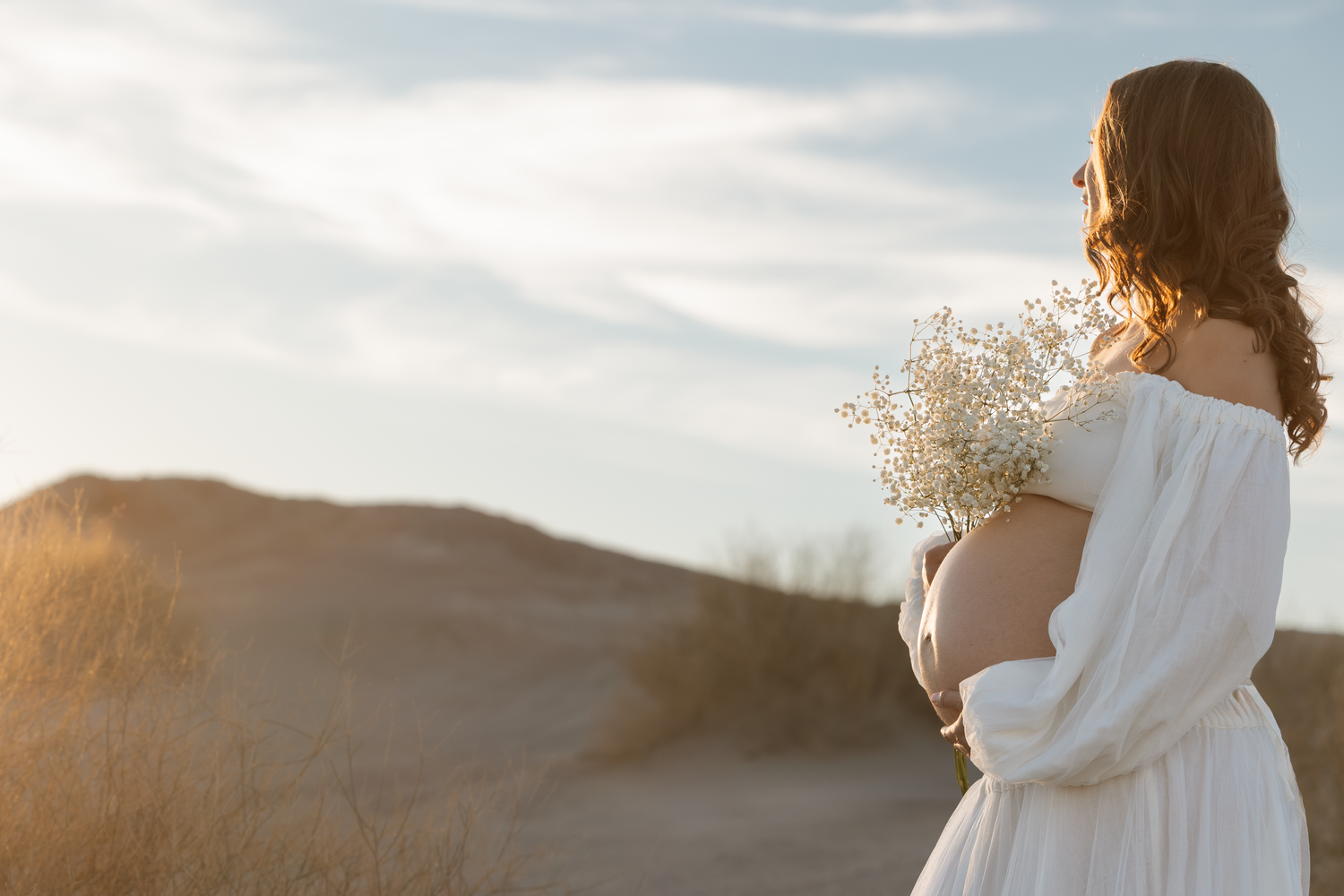 A figure in a white dress holds delicate baby's breath flowers while gazing at a sunset over rolling hills.