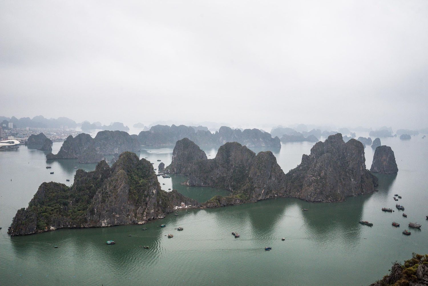Dramatic limestone karsts rise from misty waters of Ha Long Bay in Vietnam with boats scattered throughout the seascape.