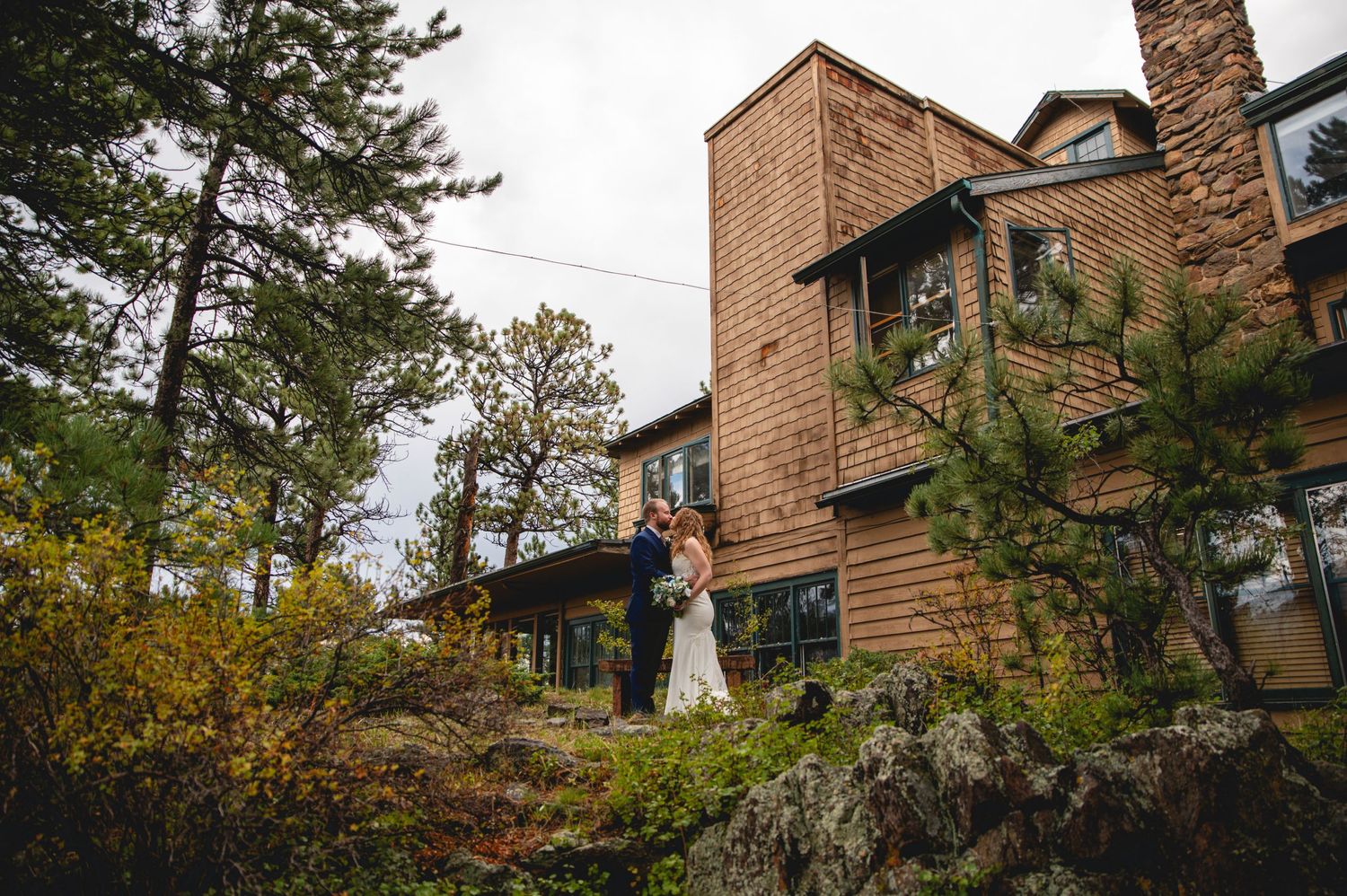 A couple poses outside a rustic wooden lodge nestled among autumn trees on a hillside.