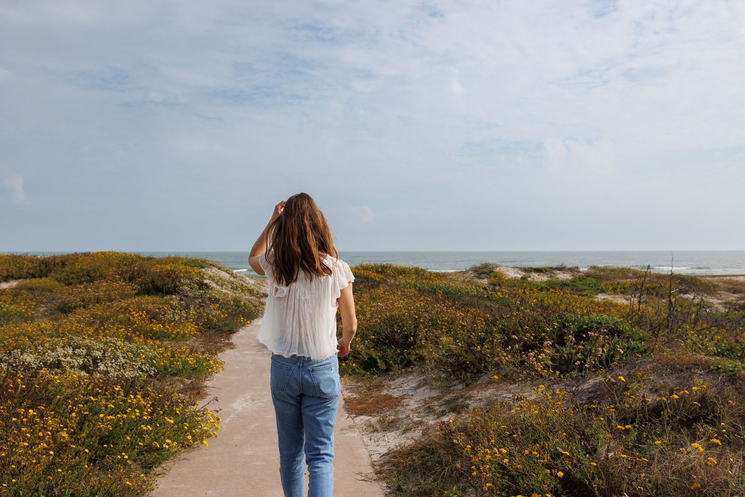 Woman in jeans walking on coastal path surrounded by wildflowers.