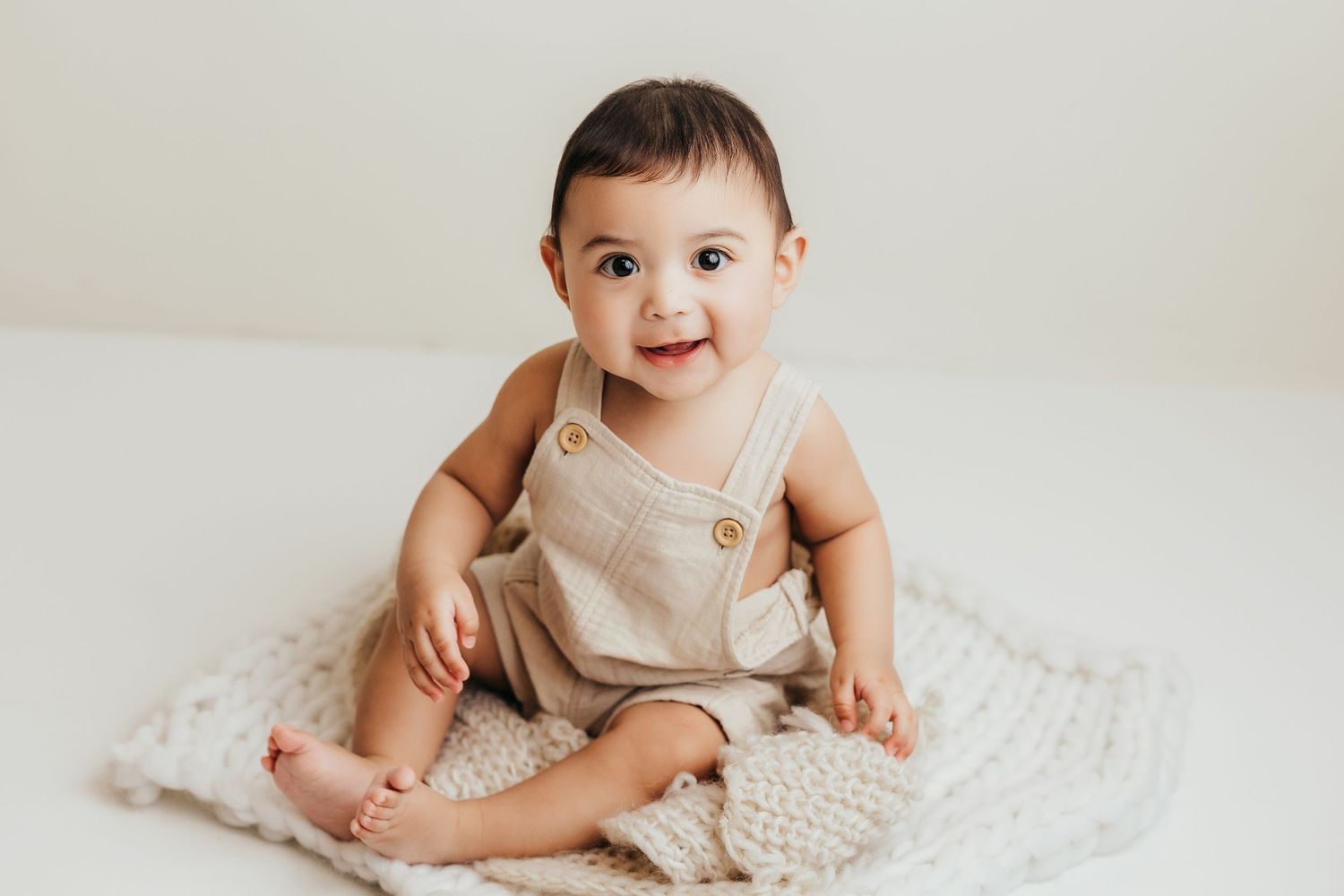 Baby in neutral-colored romper sitting and crawling on textured white blanket.