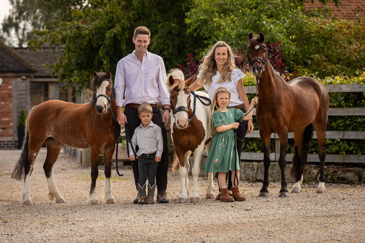 Family poses with their collection of horses of varying sizes at a stable yard.