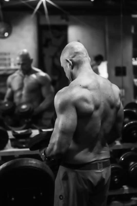 Black and white photo of bald bodybuilder displaying massive back muscles in front of gym mirror.