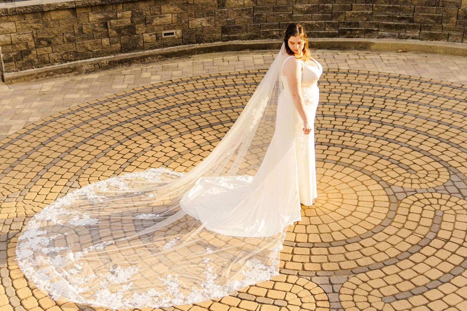 Elegant bride in a flowing white dress with long train standing on circular stone patio.