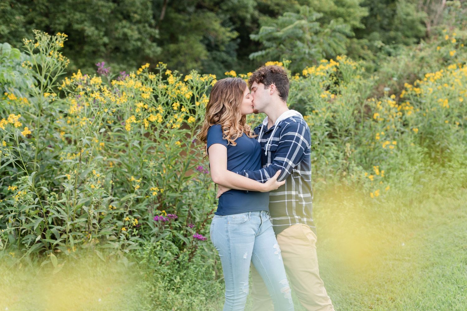 Couple walking through a sunny meadow with yellow wildflowers and green trees in the background.