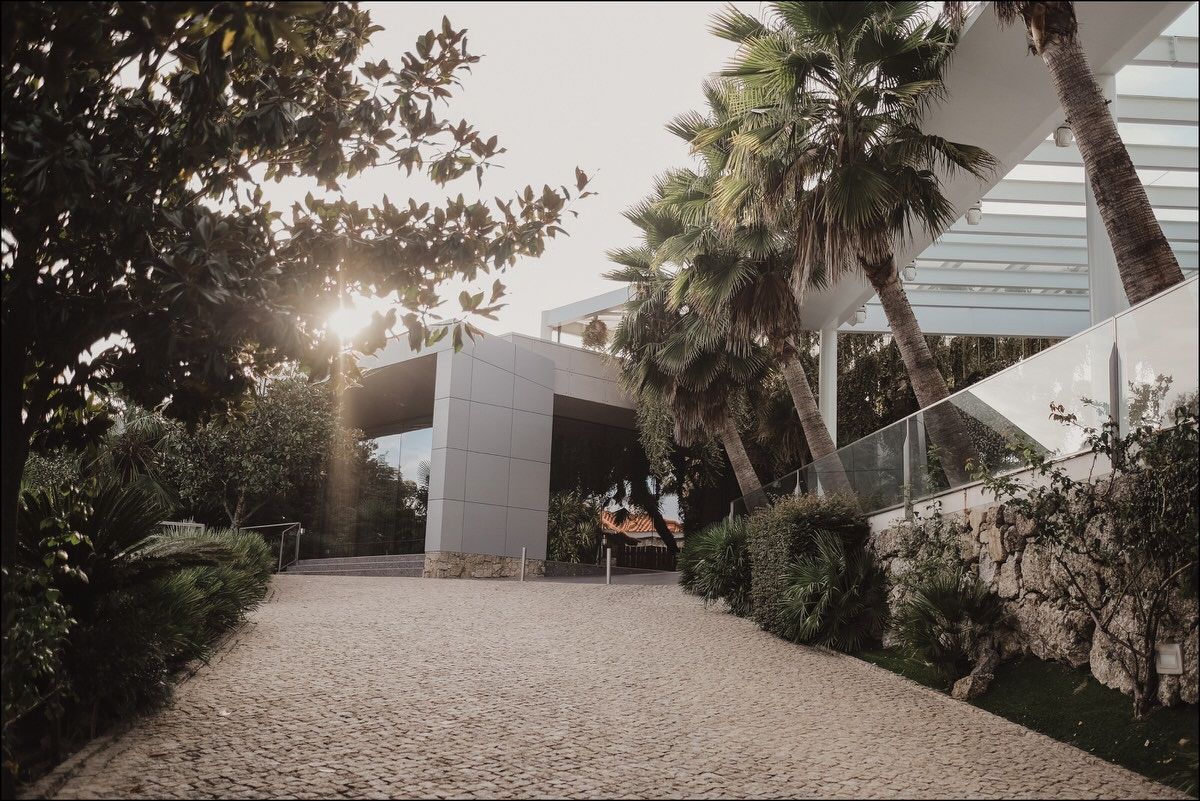 Modern white building entrance with palm trees and gravel pathway at sunset.