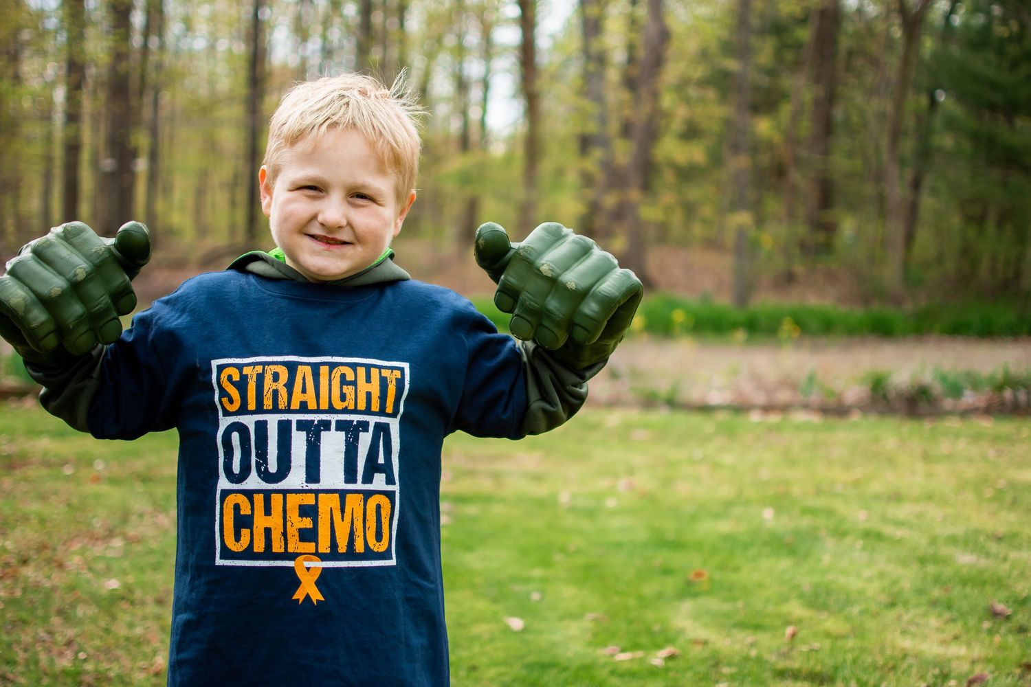 Smiling boy wearing Straight Outta Chemo shirt holds up green Hulk fists outdoors in grassy yard.