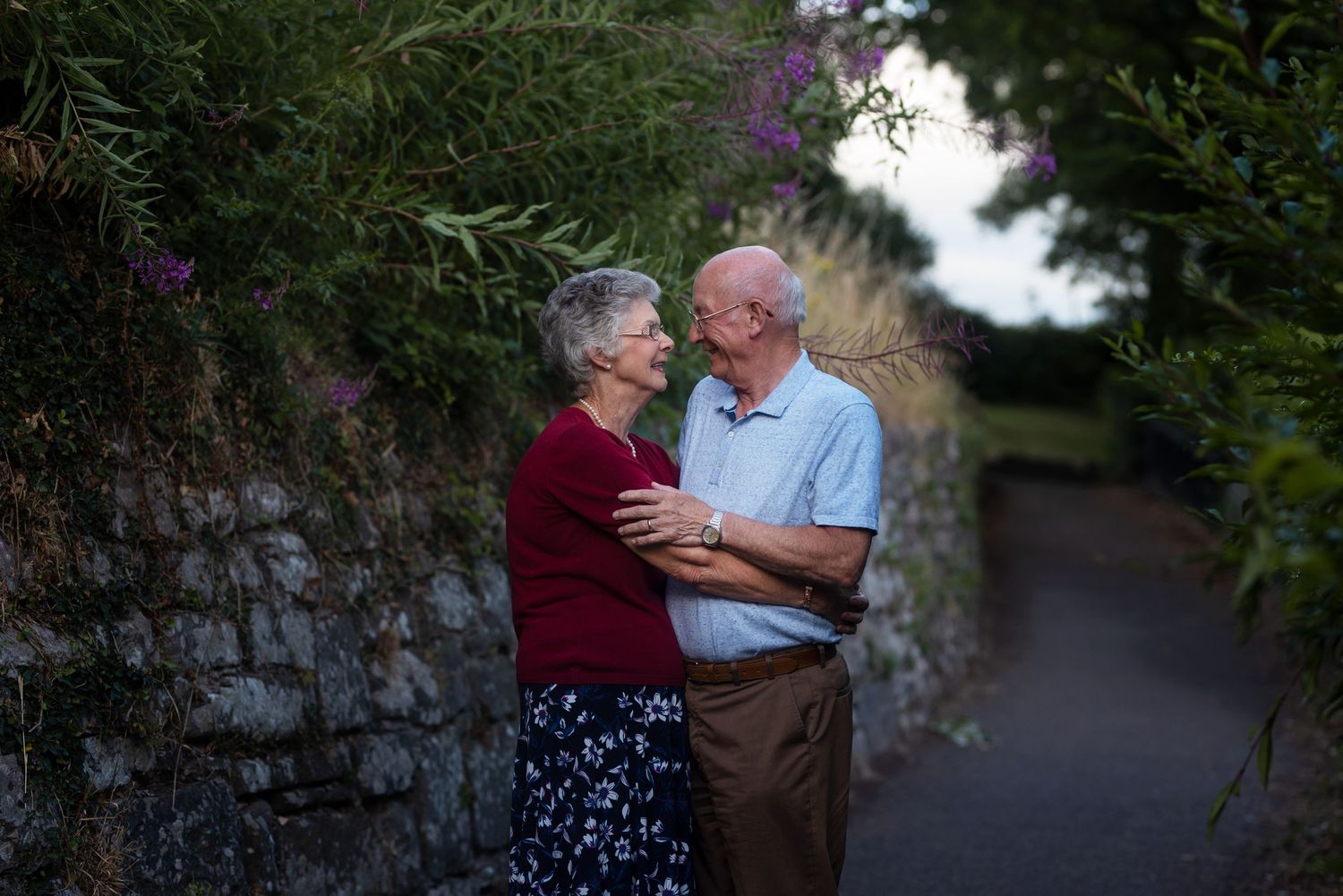 Couple sharing an intimate moment against a stone wall with purple flowers in the background.