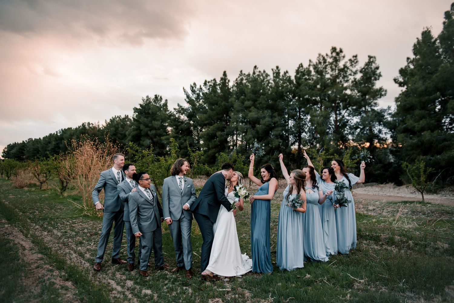 Wedding party in dusty blue dresses and gray suits pose together at sunset with trees in background.