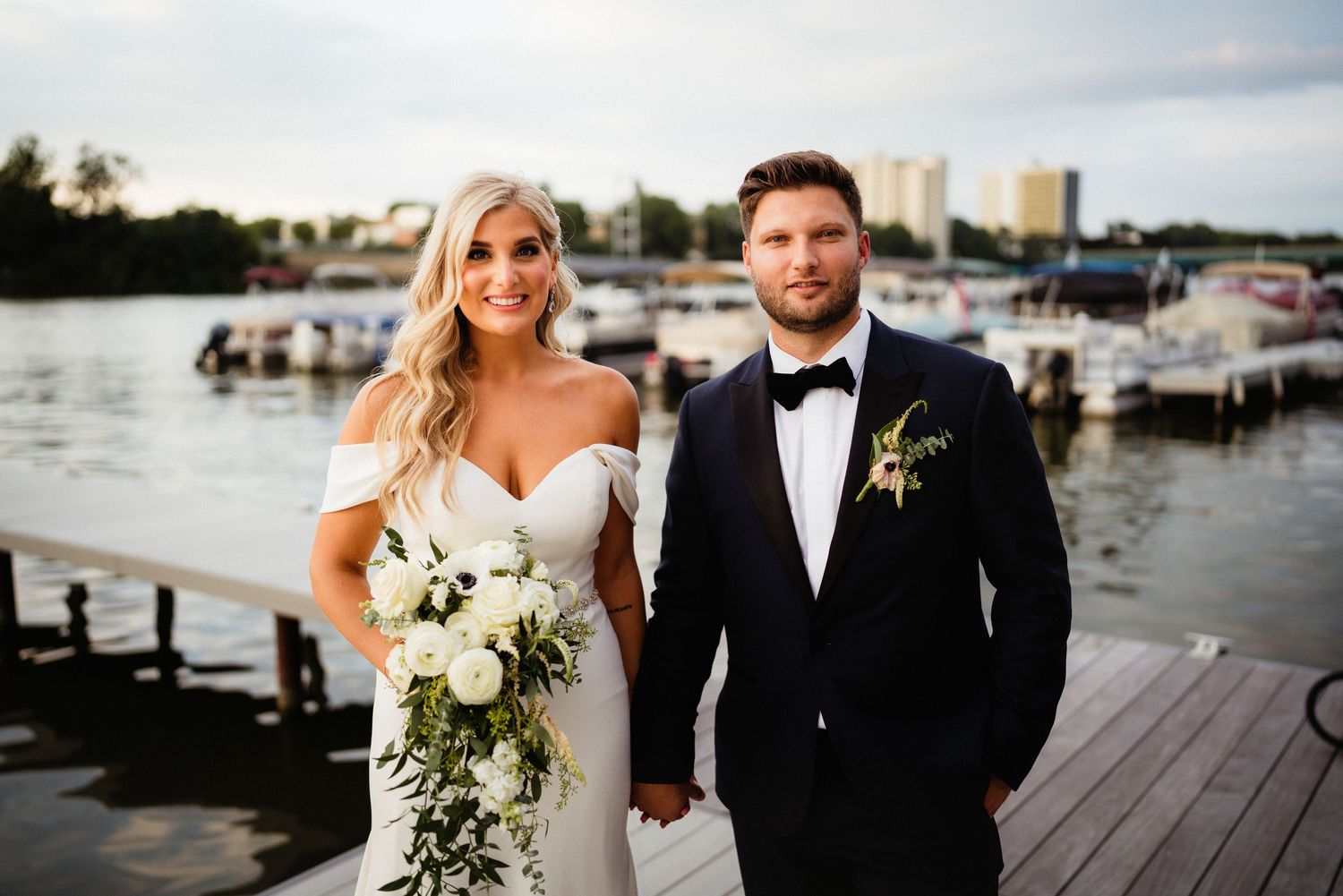 A wedding couple poses on a dock with boats and city buildings in the background at sunset.