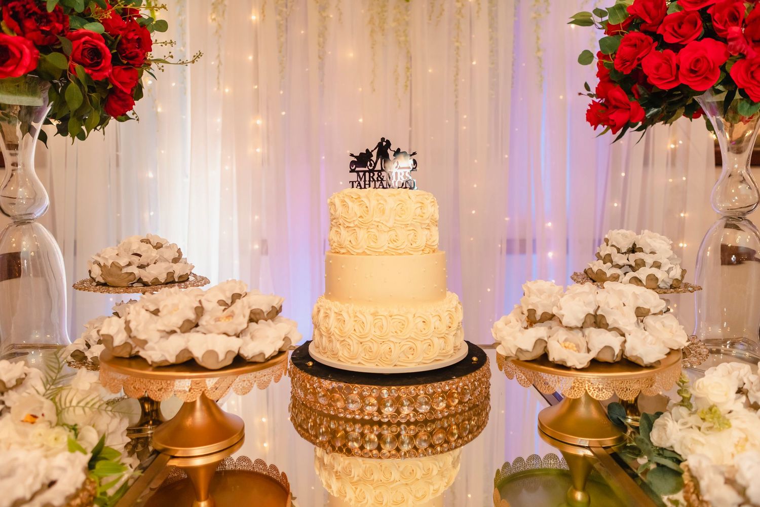 Elegant wedding dessert table featuring a tiered cream cake with gold accents and red rose floral arrangements.