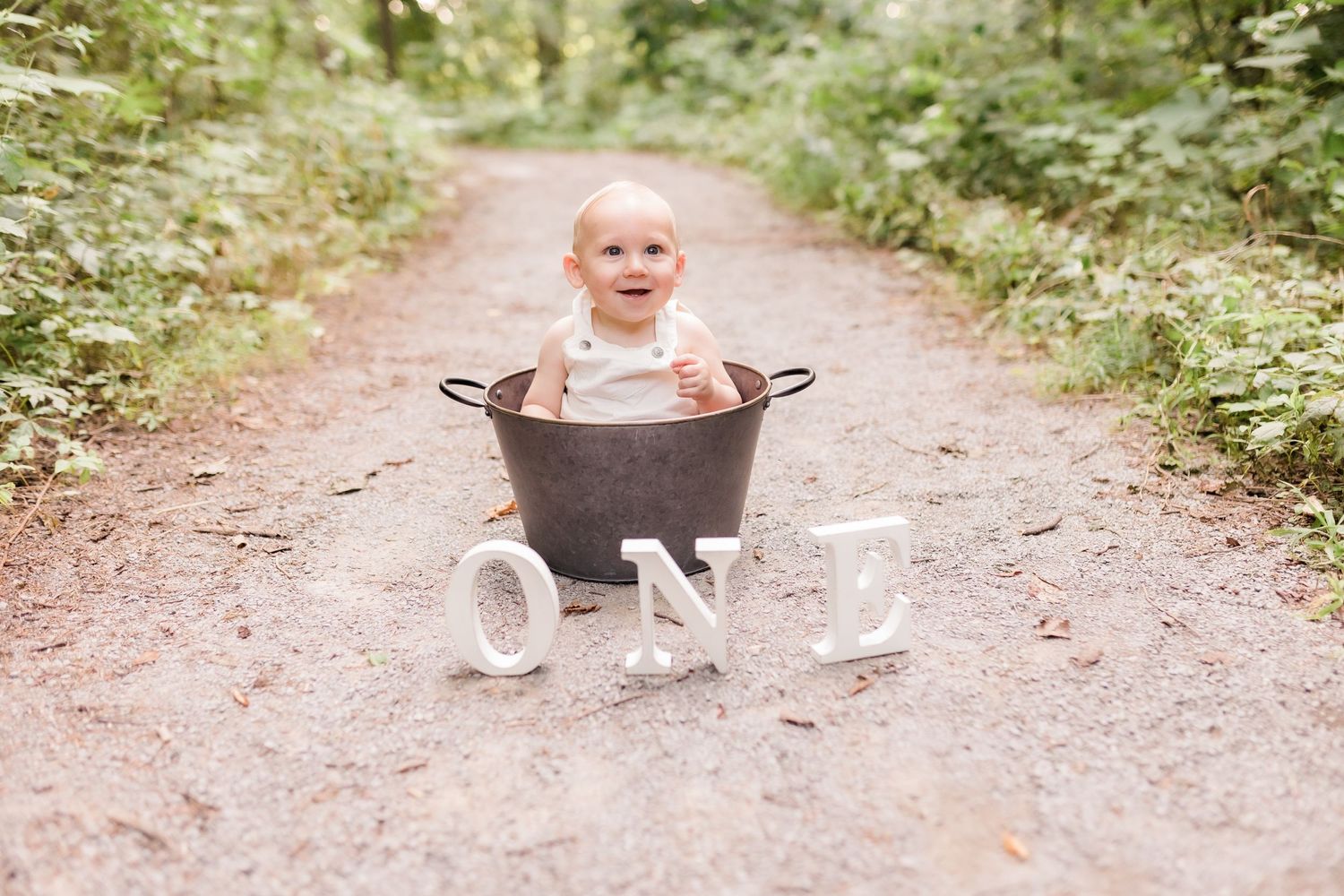 Smiling baby sitting in a vintage metal tub on a dirt path with ONE letter props for a first birthday.