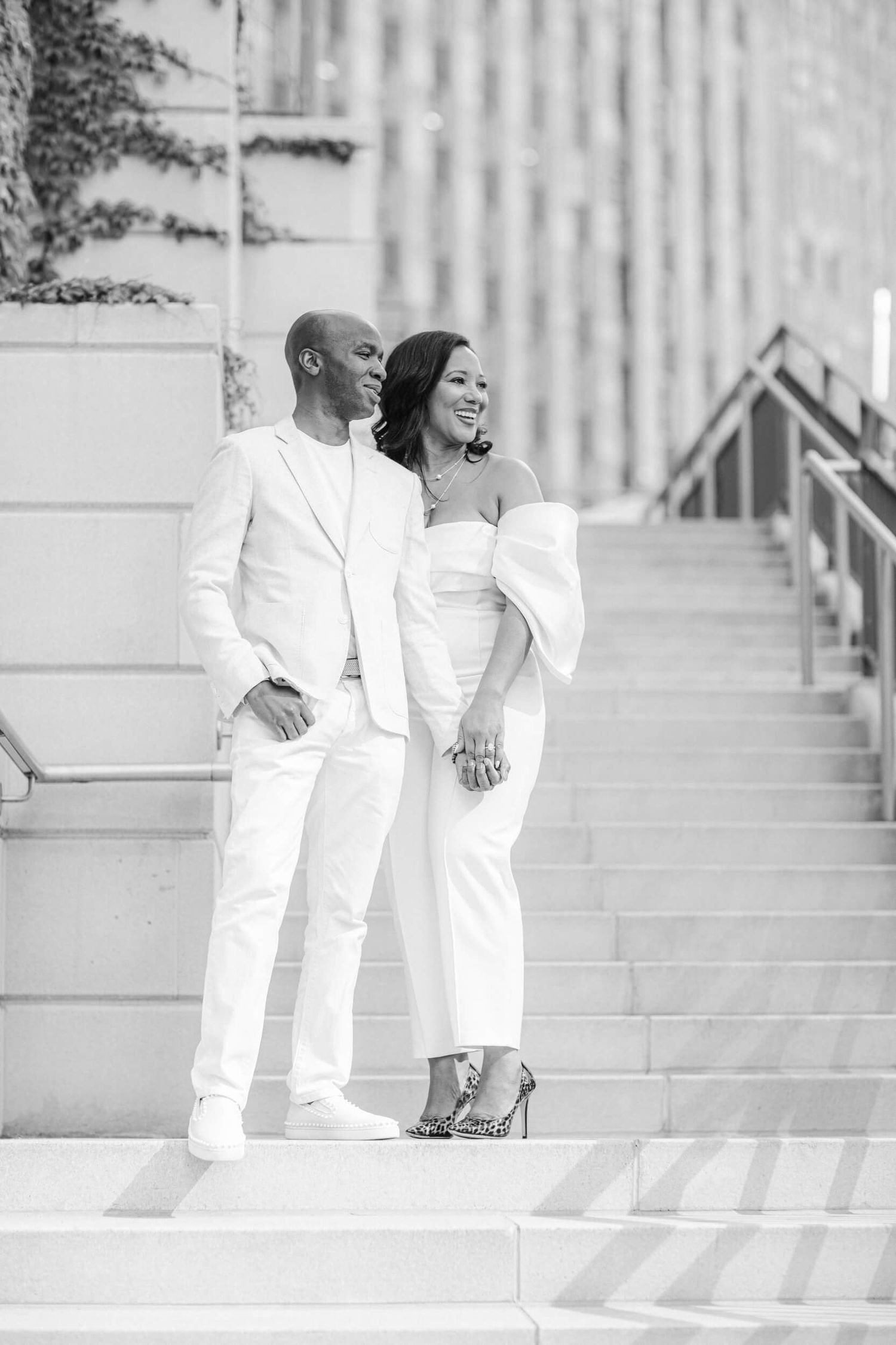 Elegant couple in all white outfits posing on architectural steps in black and white photography.