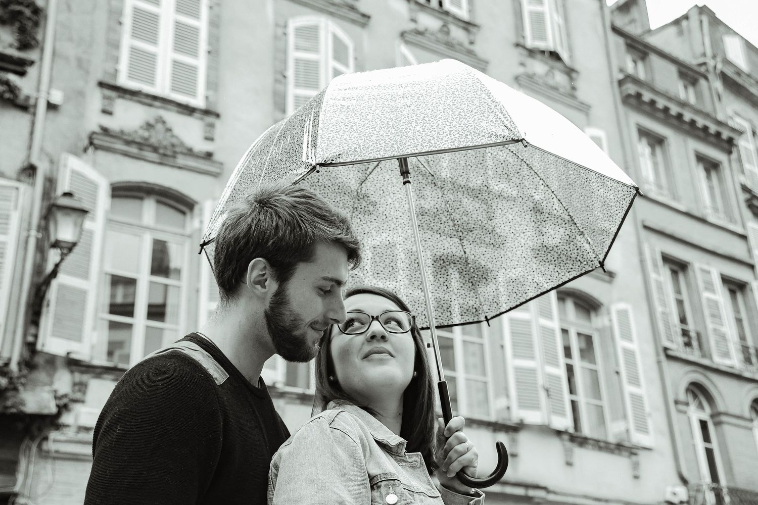 Romantic couple shares moment under clear umbrella in front of historic building.