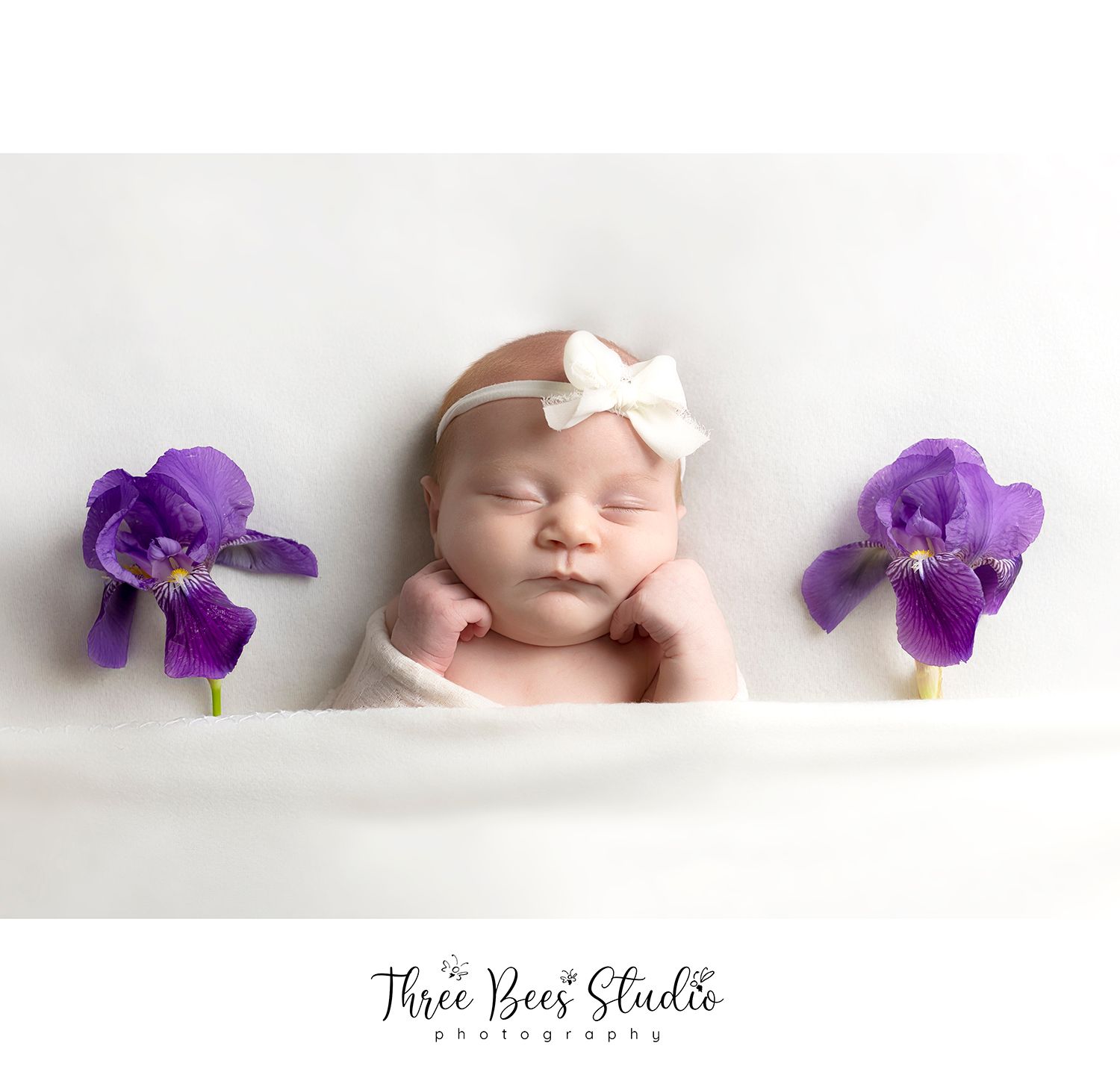 Newborn photography with a white bow headband and purple iris flowers on each side.