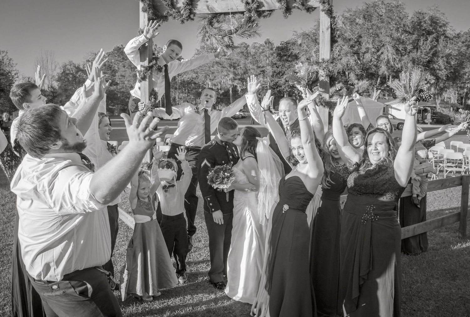 Black-and-white wedding photo of family and friends raising their hands in celebration as the newlyweds kiss.