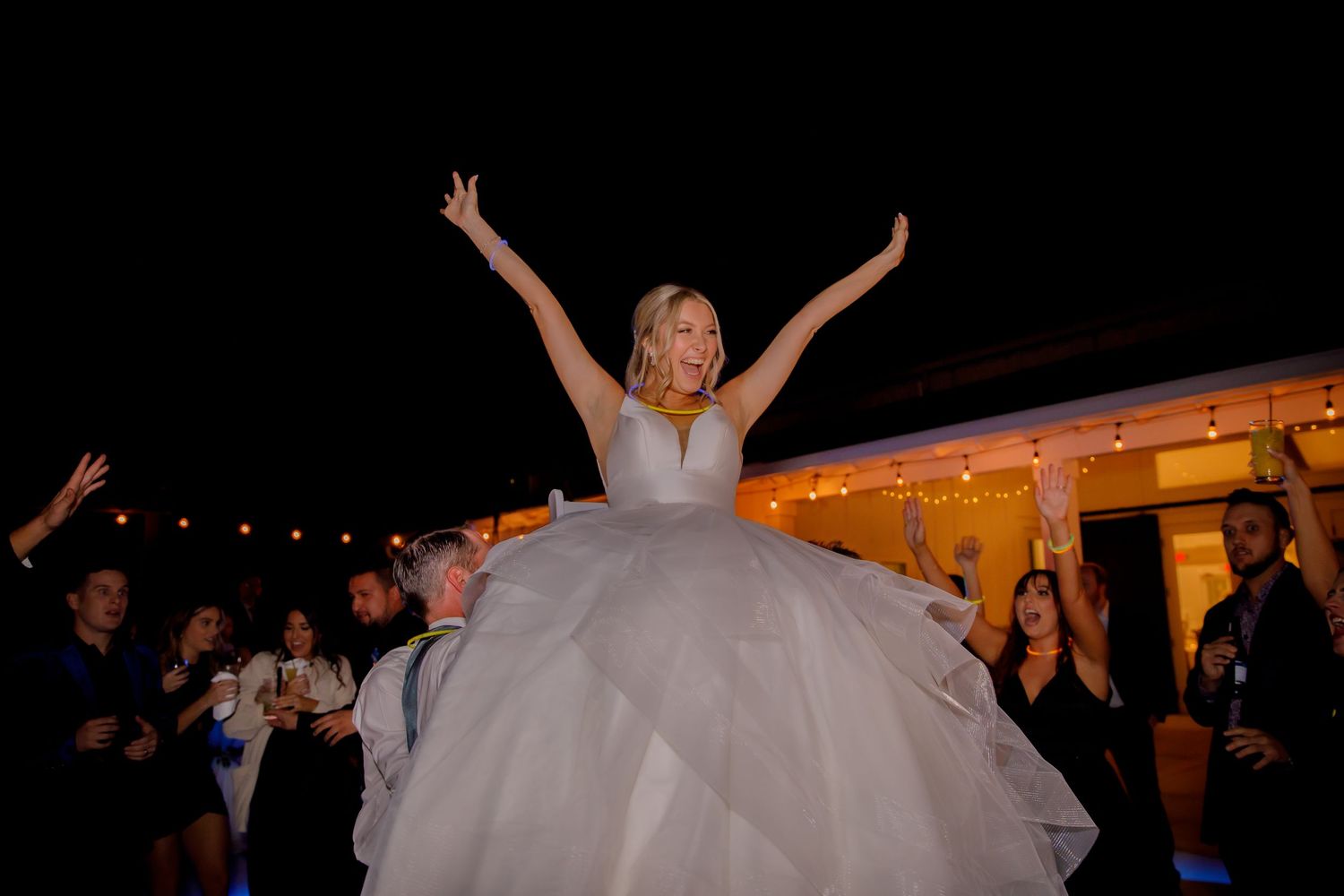 A bride in a white ballgown raises her arms in celebration during an evening reception.