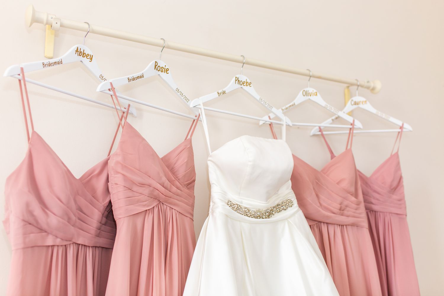 Pink bridesmaid dresses and a white wedding gown with a crystal belt hanging on white hangers against a beige wall.