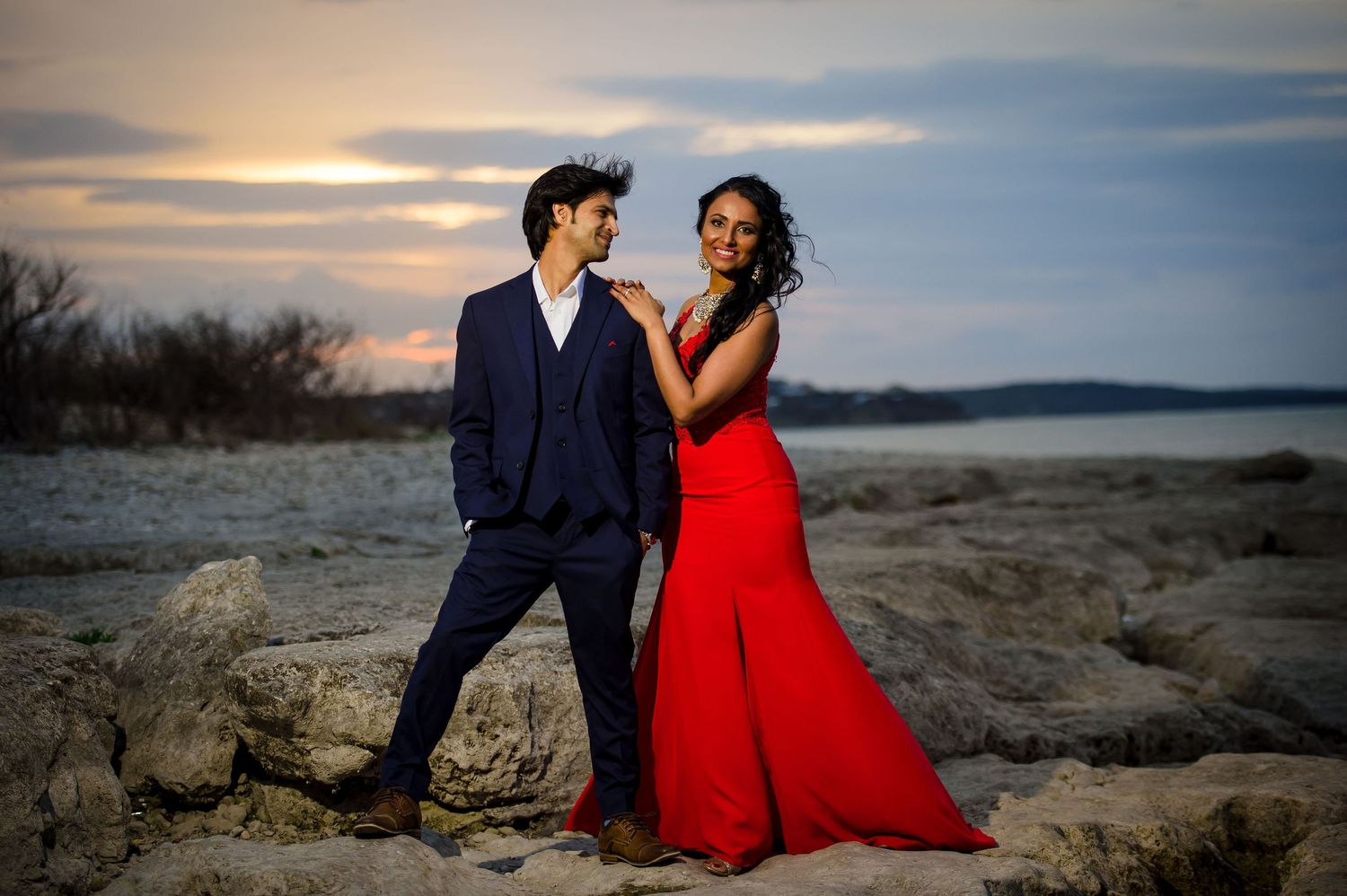 A glamorous couple poses on rocky terrain at sunset with dramatic lighting.