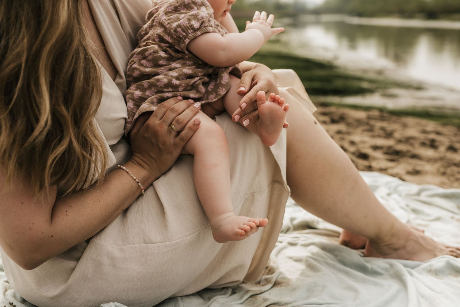 Baby's tiny feet being held while sitting by the water on a blanket.