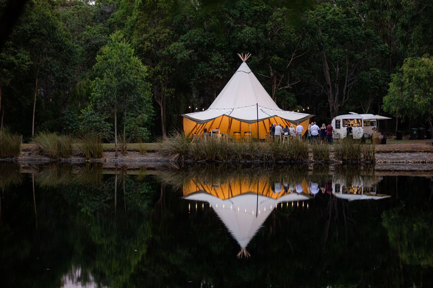 White teepee tent reflected in still lake waters at dusk.
