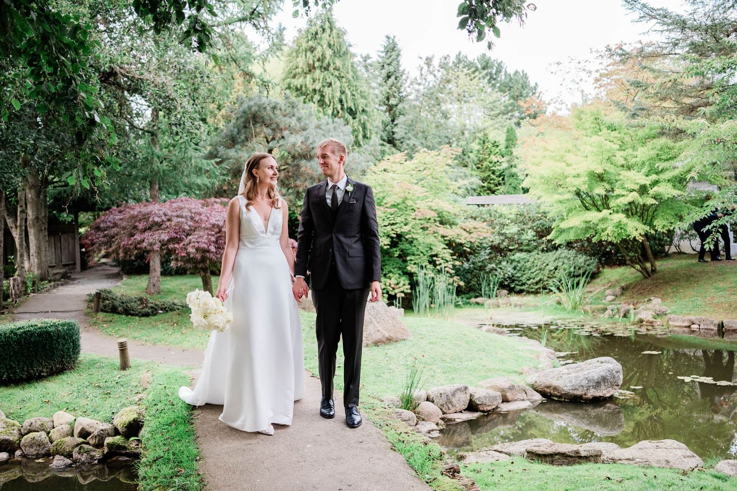 Newlyweds pose elegantly on a garden path beside a serene pond.