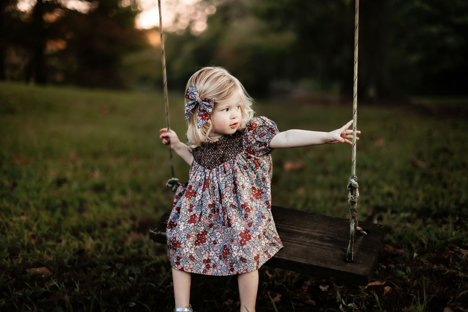 Child playing on a wooden swing in a floral dress during golden hour.